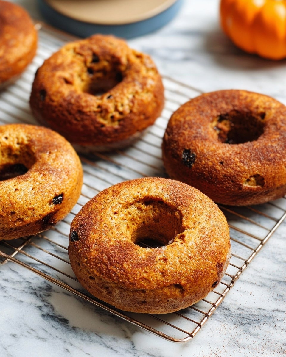 The image shows four baked donuts with a rough, golden-brown textured surface, each sprinkled with a light dusting of cinnamon or spice powder. The donuts are evenly spaced on a silver cooling rack, which sits on a white marbled surface. In the blurred background, there is a small orange pumpkin on the right side, adding a touch of autumn color. The front donut is in sharp focus, showing its soft dough texture and slightly uneven shapes, while the others gradually blur as they recede. The lighting is natural and soft, emphasizing the warm tones and texture of the donuts. Photo taken with an iphone --ar 4:5 --v 7