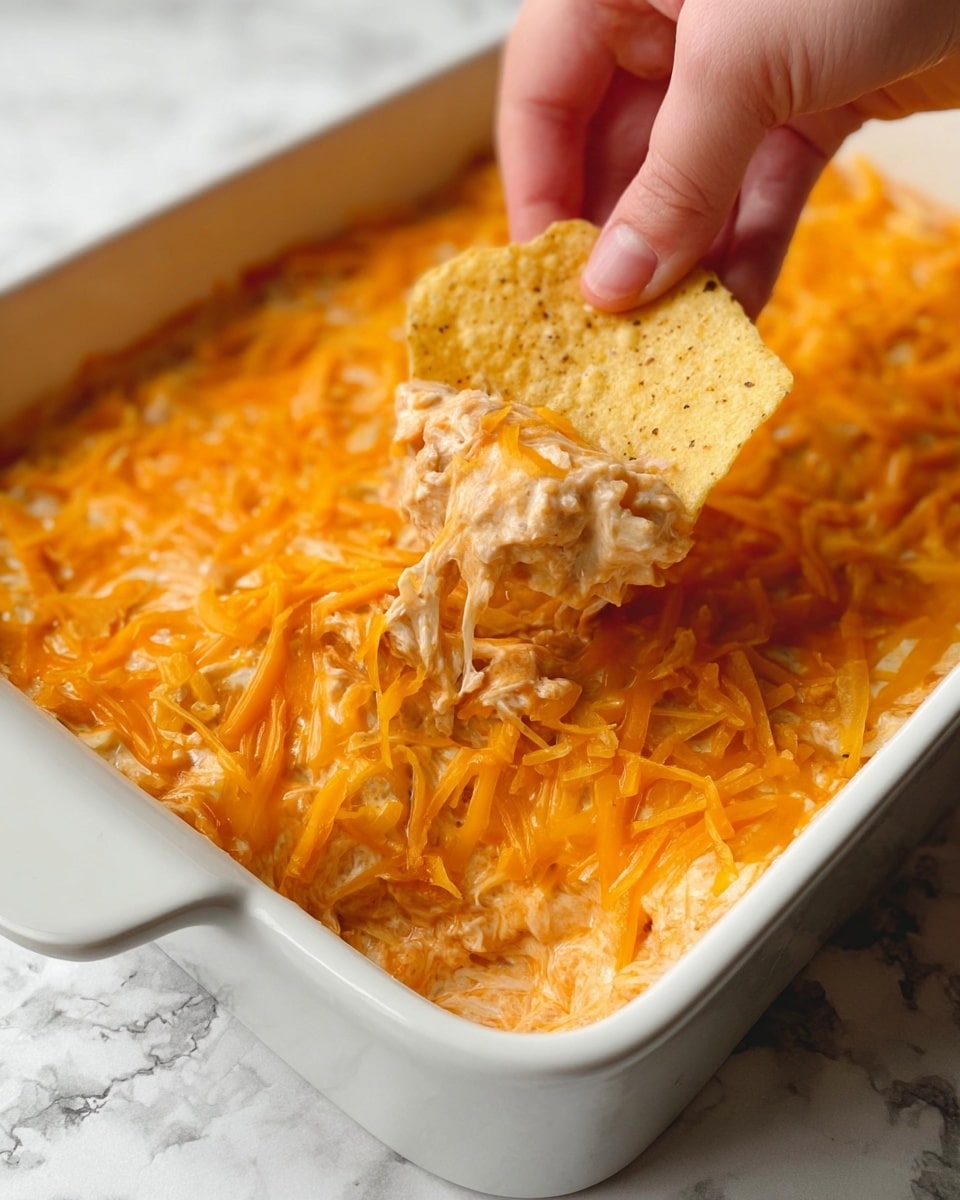 The image shows a white square baking dish filled with a layered cheesy dip. The bottom thick layer is light orange and creamy with a soft texture, likely melted cheese mixed with other ingredients. On top of it, a generous layer of shredded bright orange cheddar cheese is melted unevenly, creating a rich, gooey finish with some parts browned slightly. A woman's hand is dipping a speckled yellow tortilla chip into the dip, lifting up a scoop that reveals a chunky mix of melted cheese in shades of pale cream and orange. The dish sits on a white marbled textured surface, adding to the clean and fresh feel of the photo taken with an iphone --ar 4:5 --v 7
