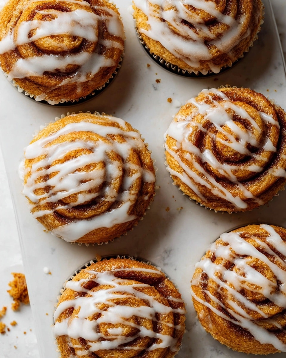 The image shows a close-up of five golden-brown cinnamon roll cupcakes inside a white muffin tray, each topped with white icing drizzled in spirals that follow the swirl pattern of the cupcake layers. The cinnamon rolls have a slightly rough texture with visible darker cinnamon stripes curling from the center outward, creating three to four spiral layers. The tray rests on a white marbled surface scattered with a few crumbs and a light dusting of cinnamon powder. Photo taken with an iphone --ar 4:5 --v 7