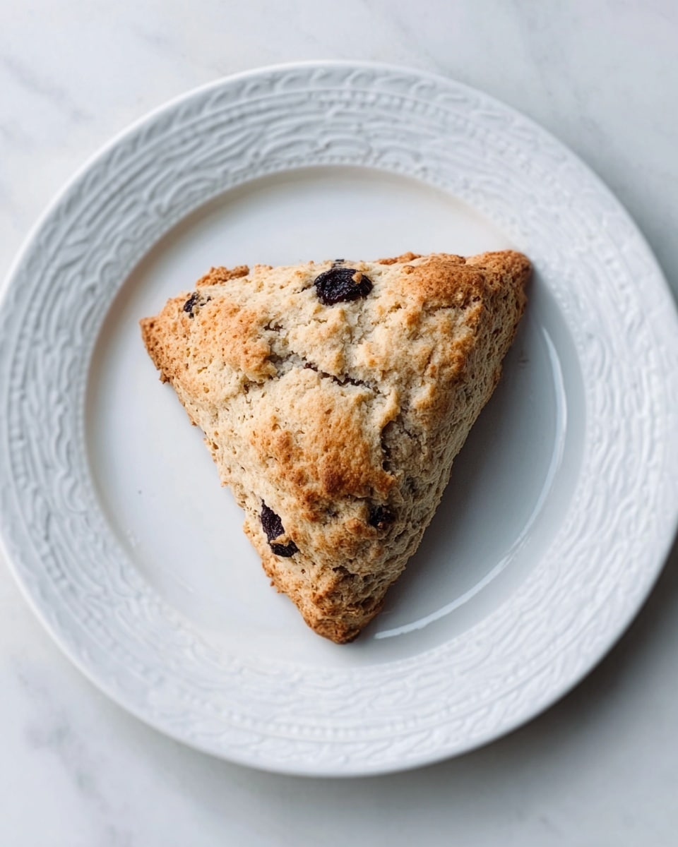 A single triangular scone with a rough, crumbly texture sits in the center of a white plate with a delicate raised pattern around the edge. The scone is light brown with darker spots where chocolate chips or berries peek through its surface. The plate is placed on a white marbled textured surface, giving a clean and simple background. The scone looks fresh and slightly crunchy on the outside, with an inviting homemade appearance. Photo taken with an iphone --ar 4:5 --v 7