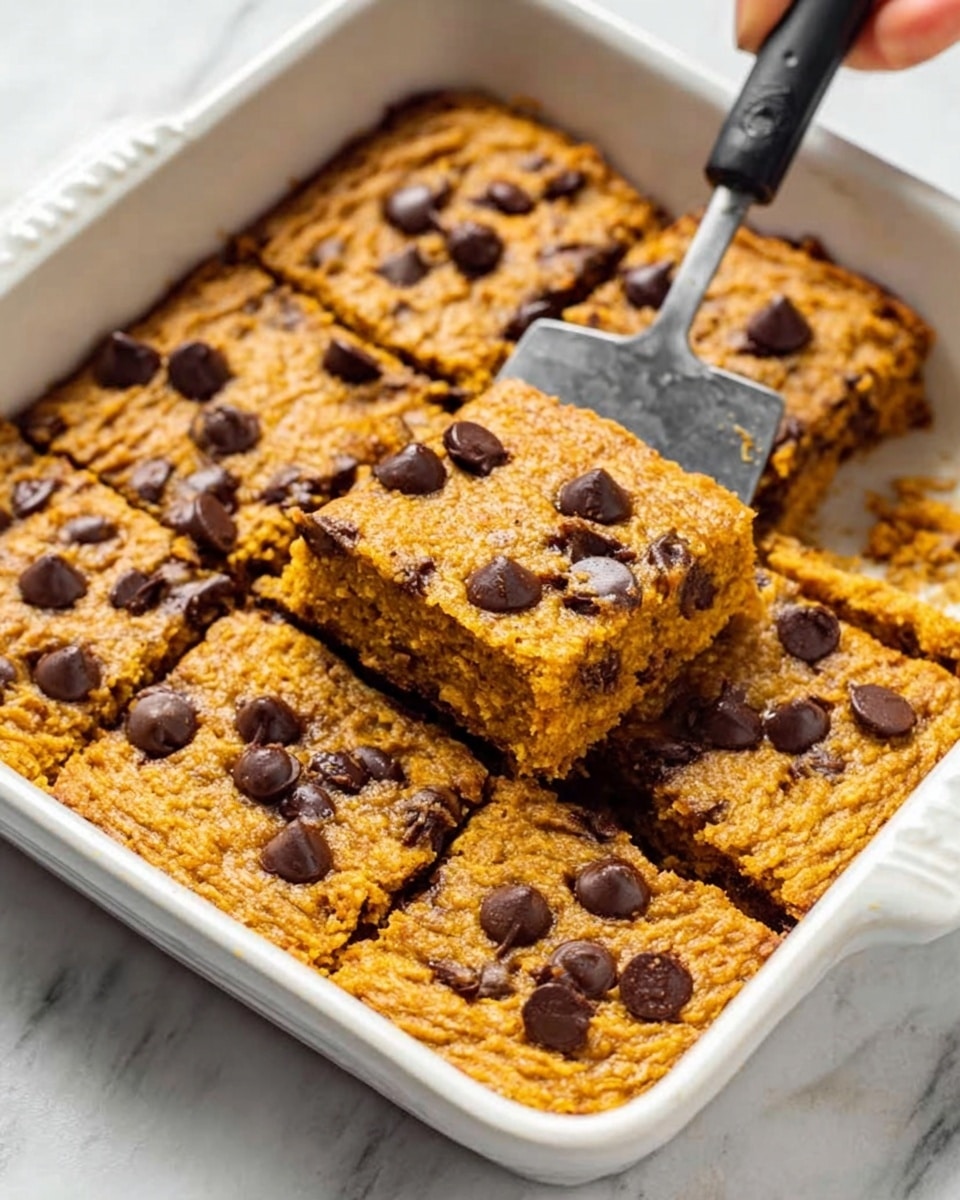 A white rectangular dish filled with a thick, golden-brown baked treat topped with scattered dark chocolate chips. A slice is cut out, showing the soft, moist inside that looks slightly dense and rich with chocolate chips inside as well. A woman’s hand holds a metal spatula lifting the cut slice from the dish. The dish is placed on a white marbled surface. Photo taken with an iphone --ar 4:5 --v 7