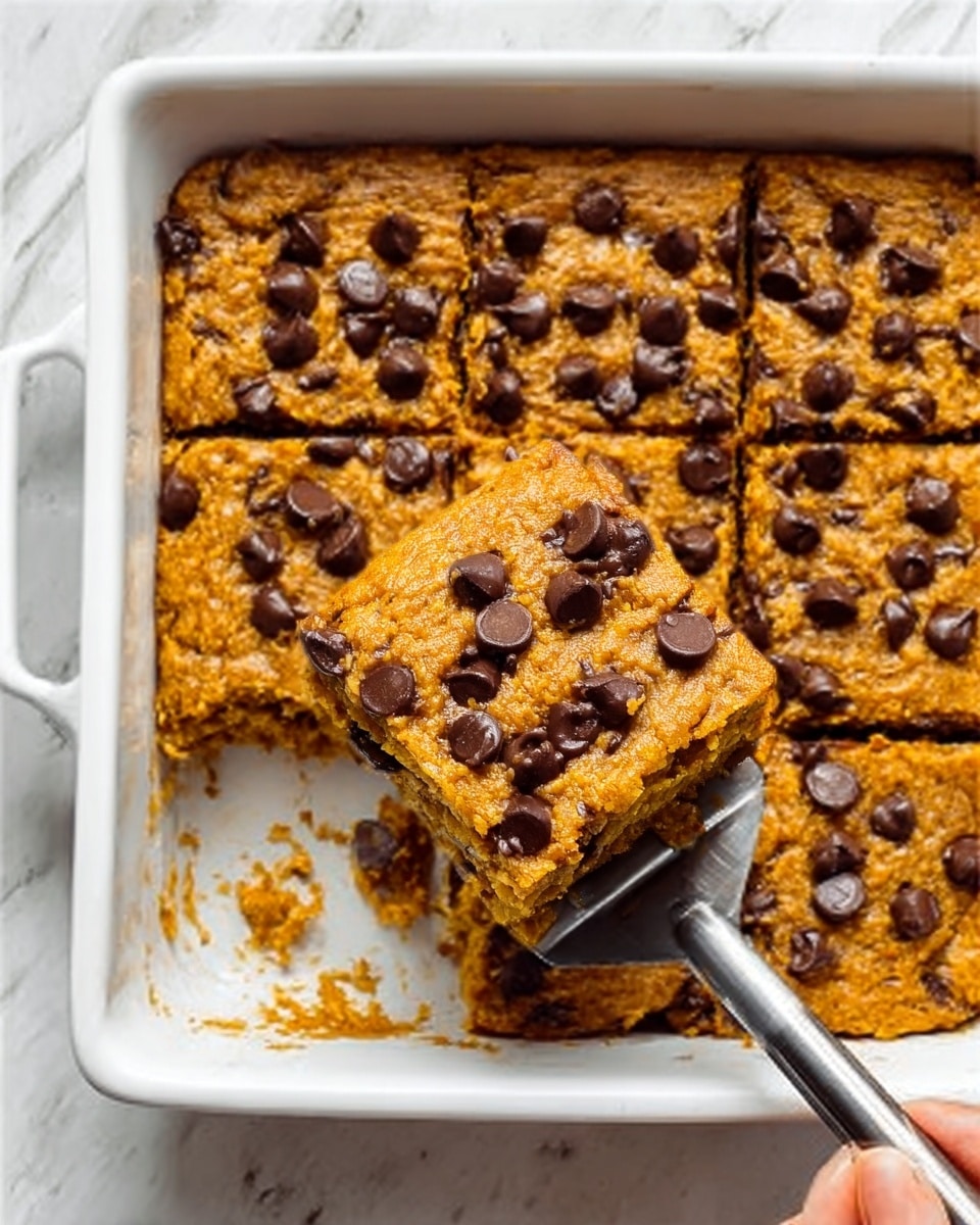 A close-up shot of a white rectangular baking dish filled with a baked pumpkin chocolate chip dessert, cut into squares. The dessert has a soft orange color with a slightly rough texture on top, dotted with shiny dark chocolate chips spread evenly across the surface. One square is lifted by a metal spatula with a black handle, showing the moist, dense inside of the dessert. A woman's hand is gently holding the spatula from the side. The dish is placed on a white marbled surface. Photo taken with an iphone --ar 4:5 --v 7