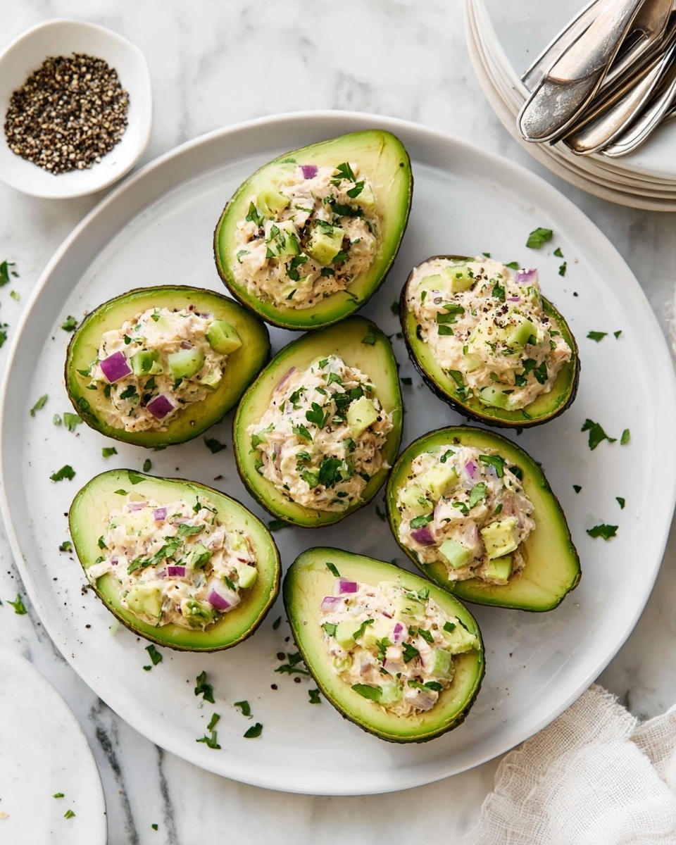 The image shows halved avocados with dark green, bumpy skin and light green smooth flesh, each filled with a creamy mixture of tuna salad that contains chunks of light pink tuna, small pieces of purple onion, green celery bits, and specks of green herbs mixed throughout. These avocado halves are arranged on a large, round white plate on a white marbled surface. Fresh green parsley leaves are placed in the foreground outside the plate, adding a touch of brightness and freshness. The mix inside the avocados looks moist and slightly chunky, contrasting with the smooth, firm avocado base. photo taken with an iphone --ar 4:5 --v 7