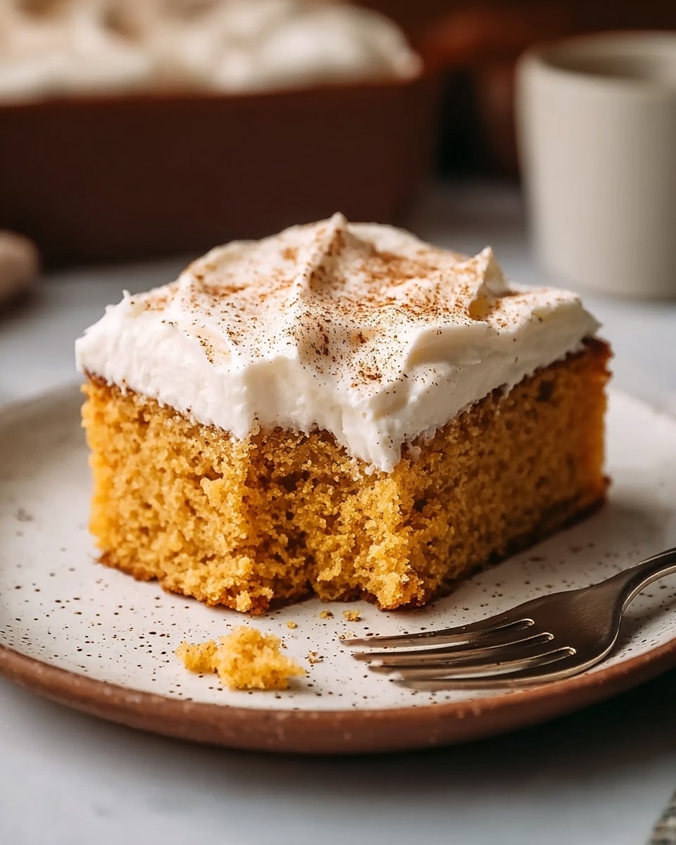 A single square piece of moist orange-brown cake sits on a white plate with a textured rim, showing a crumbly and soft texture. On top of the cake, there is a thick, creamy white frosting spread unevenly with small peaks and sprinkled with fine brown powder. To the right of the cake, a silver fork rests on the plate, with a few crumbs scattered nearby. The background has a soft blur with warm tones and a white marbled texture surface underneath the plate. photo taken with an iphone --ar 4:5 --v 7