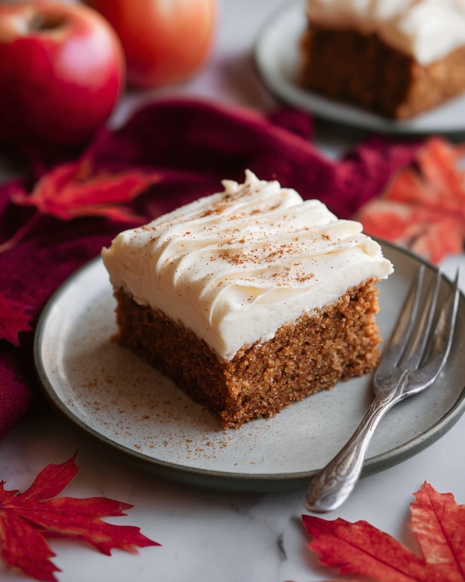 A square piece of moist brown cake with a crumbly texture forms the base layer, topped with a thick, smooth, white frosting layer that has soft, wavy ridges and a light sprinkling of brown spice on top, all placed on a white plate with a matte finish. The plate sits on a white marbled surface, surrounded by red autumn leaves, a silver fork next to the cake, a red apple in the background, and a deep red cloth nearby. Photo taken with an iphone --ar 4:5 --v 7