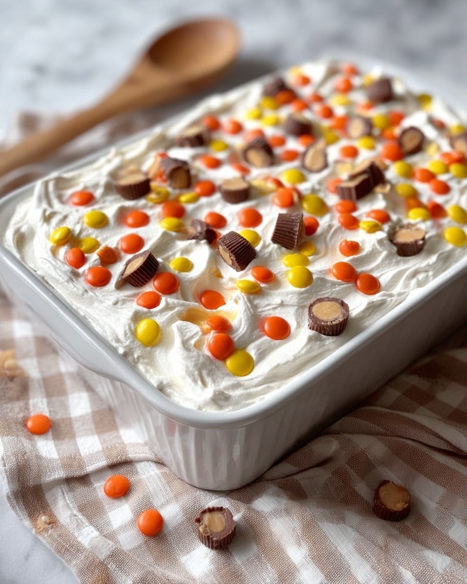 A white deep rectangular dish filled with a thick white whipped cream layer that looks soft and smooth on top. Scattered all over the whipped cream layer are small colorful candy pieces in bright orange, yellow, and brown, along with mini chocolate peanut butter cup candies that are cut into halves and some whole, adding texture and color contrast. The dish sits on a cloth with a soft checkered pattern in light beige on a white marbled surface, and a wooden spoon is blurred in the background. The overall look is inviting and festive, with a mix of creamy and crunchy textures. photo taken with an iphone --ar 4:5 --v 7