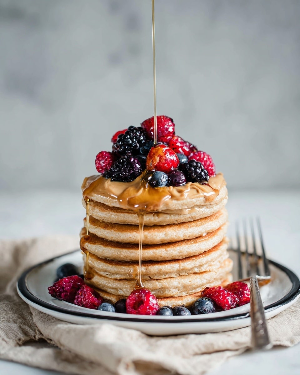 A stack of six light brown pancakes with a soft, fluffy texture is centered on a white plate with a black rim, which rests on a beige cloth over a white marbled surface. On top, there is a layer of smooth peanut butter spreading across the pancake surface, holding a mix of fresh berries: red raspberries, dark blackberries, and deep blue blueberries. Golden syrup is being poured over the berries, creating shiny, dripping streams that flow down the sides of the pancake stack. Some berries are also placed around the base of the stack on the plate. To the right of the plate, a silver fork lies on the cloth, slightly out of focus. The background is a plain, soft gray, adding contrast to the vibrant colors of the food. photo taken with an iphone --ar 4:5 --v 7