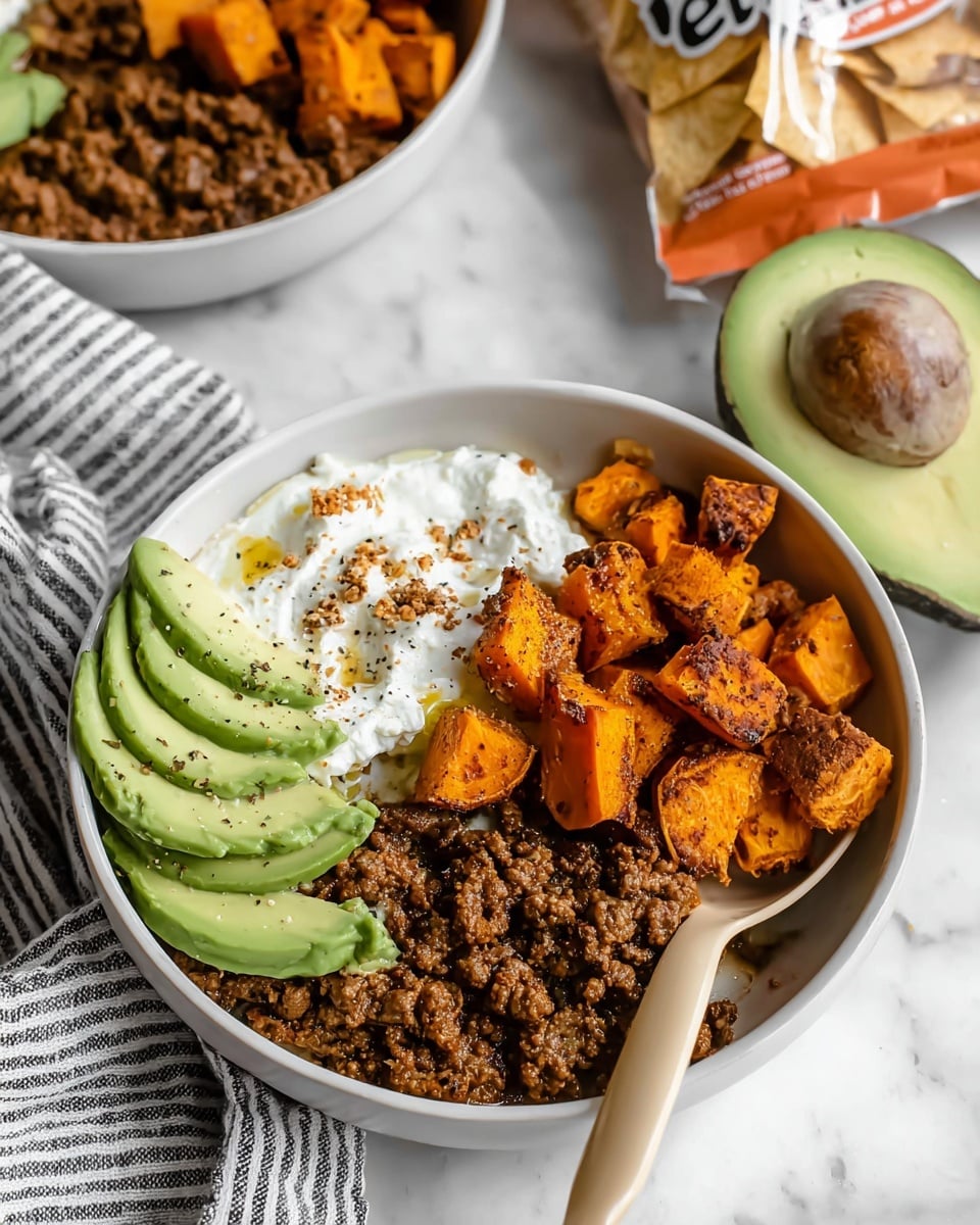The image shows a white bowl with three main layers: a pile of cooked ground meat with a crumbly texture in dark brown color on the bottom right, next to roasted sweet potato cubes in bright orange with slightly charred edges at the top, and a creamy white layer of cottage cheese with a few drops of sauce and a sprinkle of black pepper on the left. Thin slices of fresh avocado in light green are placed on top of the meat near the center, adding a smooth texture. A beige spoon rests inside the bowl on the right side. The bowl is placed on a white marbled surface, with a cut avocado half and a striped cloth near it. Part of another similar bowl and a bag of tortilla chips can be seen in the background. Photo taken with an iphone --ar 4:5 --v 7