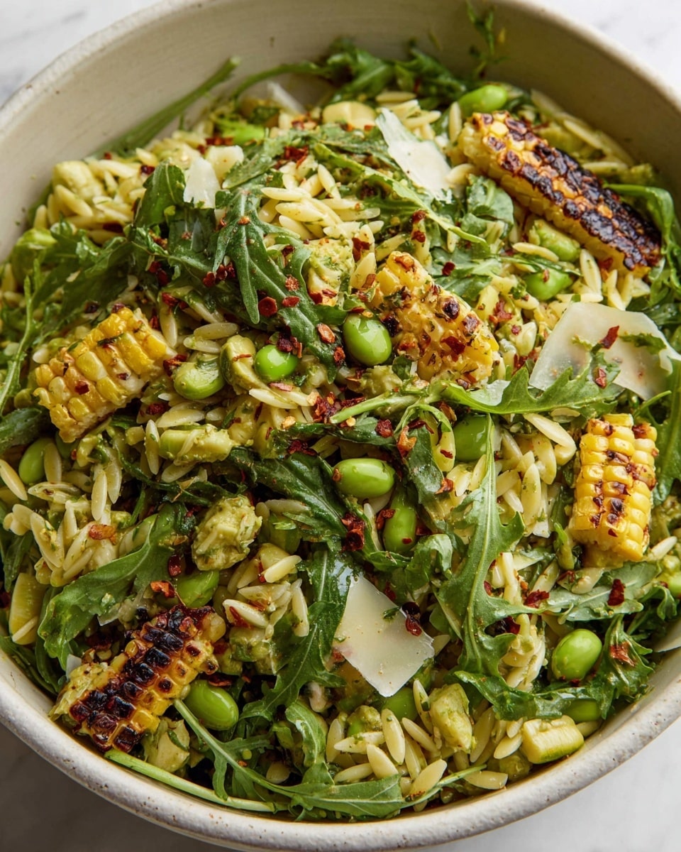 The image shows a close-up of a mixed salad served in a white bowl, placed on a white marbled surface. The salad has several layers: the base consists of light yellow orzo pasta with a soft texture, scattered throughout are dark green arugula leaves that add a leafy texture and color contrast. On top, pieces of charred yellow corn cut from the cob add a slightly blackened, grilled look. Small bright green edamame beans are mixed in, giving a smooth, rounded appearance. There are also thin slices of white shaved cheese spread around, and the entire dish is lightly sprinkled with red chili flakes and herbs, adding specks of red and green seasoning. The bowl is filled generously, showing a colorful and textured salad mix. photo taken with an iphone --ar 4:5 --v 7