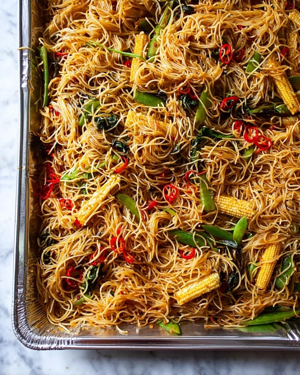 The image shows a large silver baking tray filled with stir-fried thin noodles mixed with several ingredients. The noodles are light golden with hints of brown sauce, spread unevenly across the tray. Mixed within the noodles are bright yellow baby corn pieces, small dark green leafy vegetables, green snap peas, and thin sliced red chili peppers, giving pops of red and green color. The noodles and vegetables look slightly glossy from the sauce and have a slightly tangled texture throughout. The tray is set on a white marbled surface. photo taken with an iphone --ar 4:5 --v 7