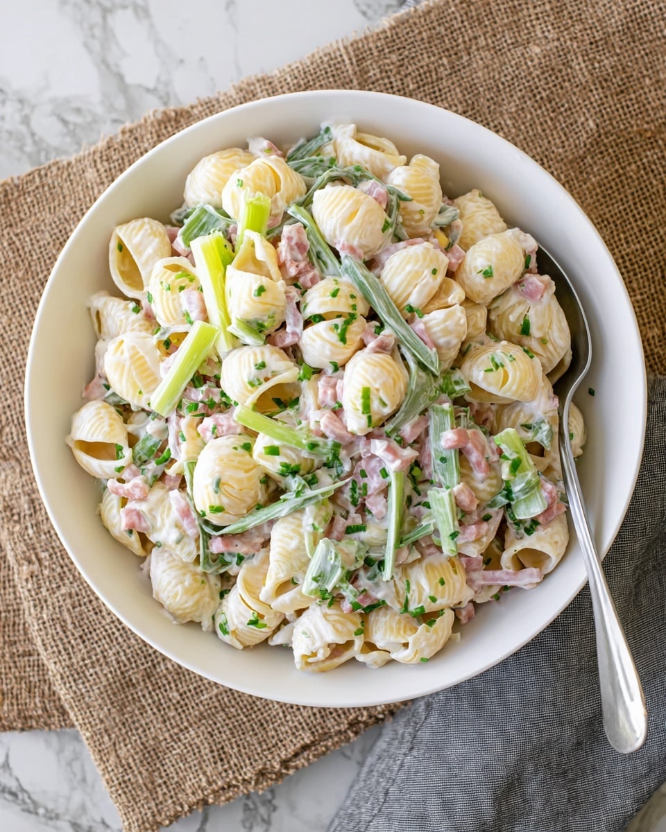 A white bowl filled with creamy pasta salad featuring two types of pasta: yellow shell-shaped pieces and flat green pasta strips. The pasta is mixed well with a white creamy sauce, small bits of diced ham, and sprinkled with chopped green herbs and celery leaves on top. There is a silver spoon resting inside the bowl to the right, and the bowl is placed on a textured burlap cloth over a white marbled surface, with a gray cloth napkin partially visible on the right side. Photo taken with an iphone --ar 4:5 --v 7
