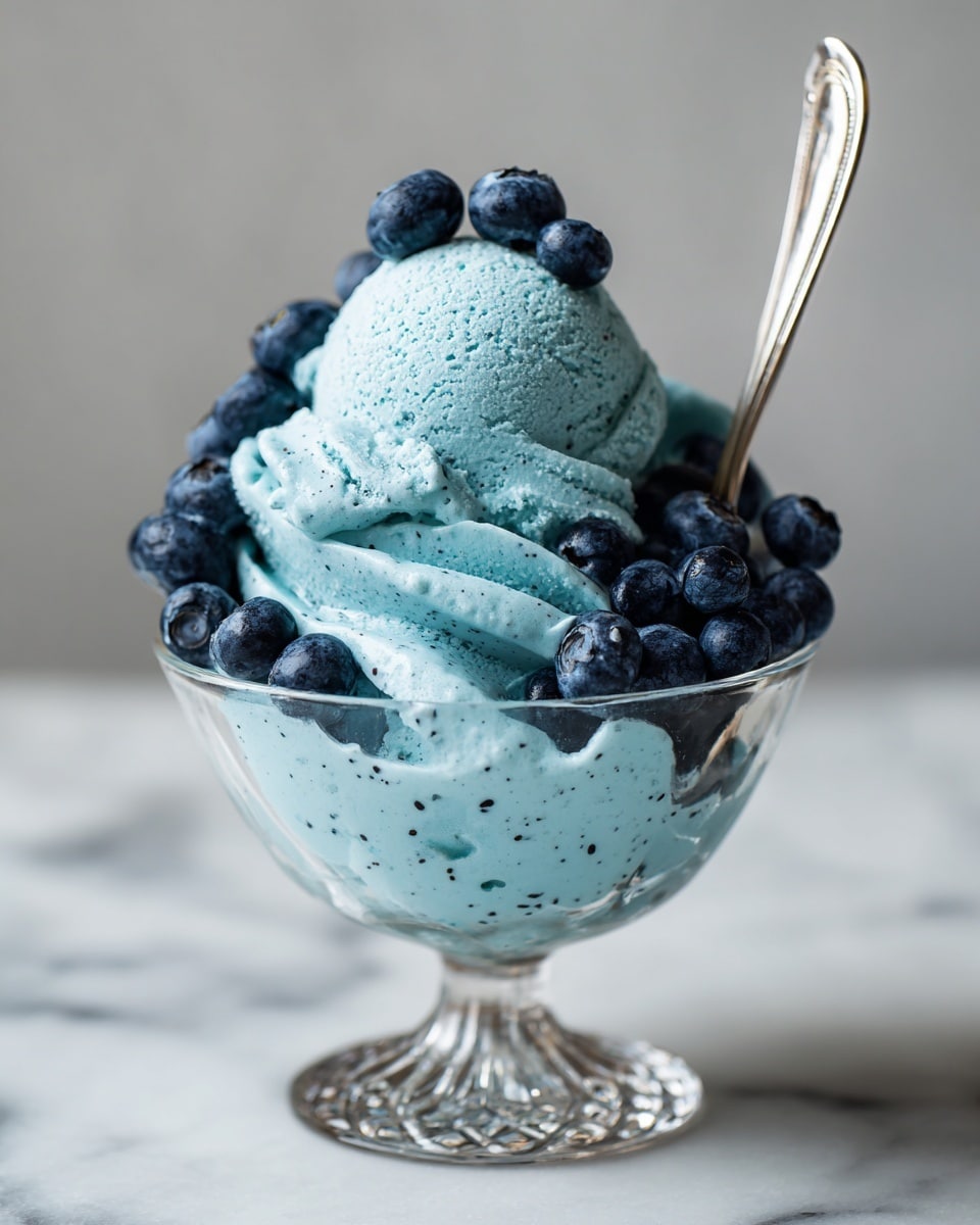 A glass dessert cup filled with creamy bright blue ice cream that has a smooth texture with tiny dark specks. The ice cream forms several thick waves around a perfect round scoop placed on top, giving a layered look with the base layer and the scoop as the second layer. Fresh dark blue blueberries are scattered on and around the ice cream, adding a pop of natural color and texture. A silver spoon is inserted into the ice cream on the right side. The cup has a shiny, patterned stem and base, sitting on a white marbled surface. Photo taken with an iphone --ar 4:5 --v 7