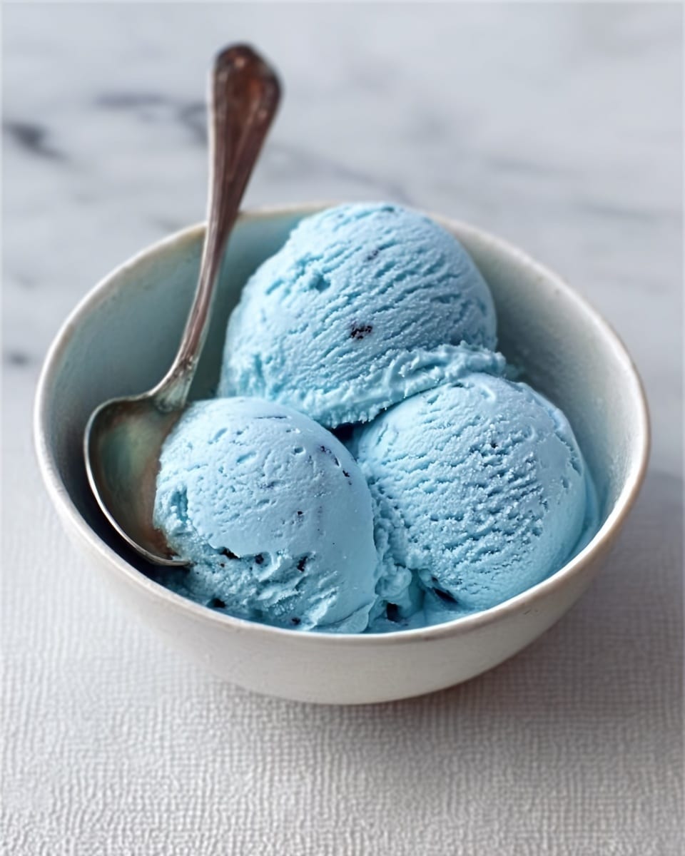The image shows a white bowl filled with three scoops of light blue ice cream, each scoop having a smooth yet slightly creamy texture with soft shadows. Inside the bowl, a silver spoon rests on the side, partially touching the ice cream. The bowl is set on a white marbled surface, providing a clean and simple background. The ice cream has a soft, cold look with small air pockets and gentle curves on each scoop. Photo taken with an iphone --ar 4:5 --v 7