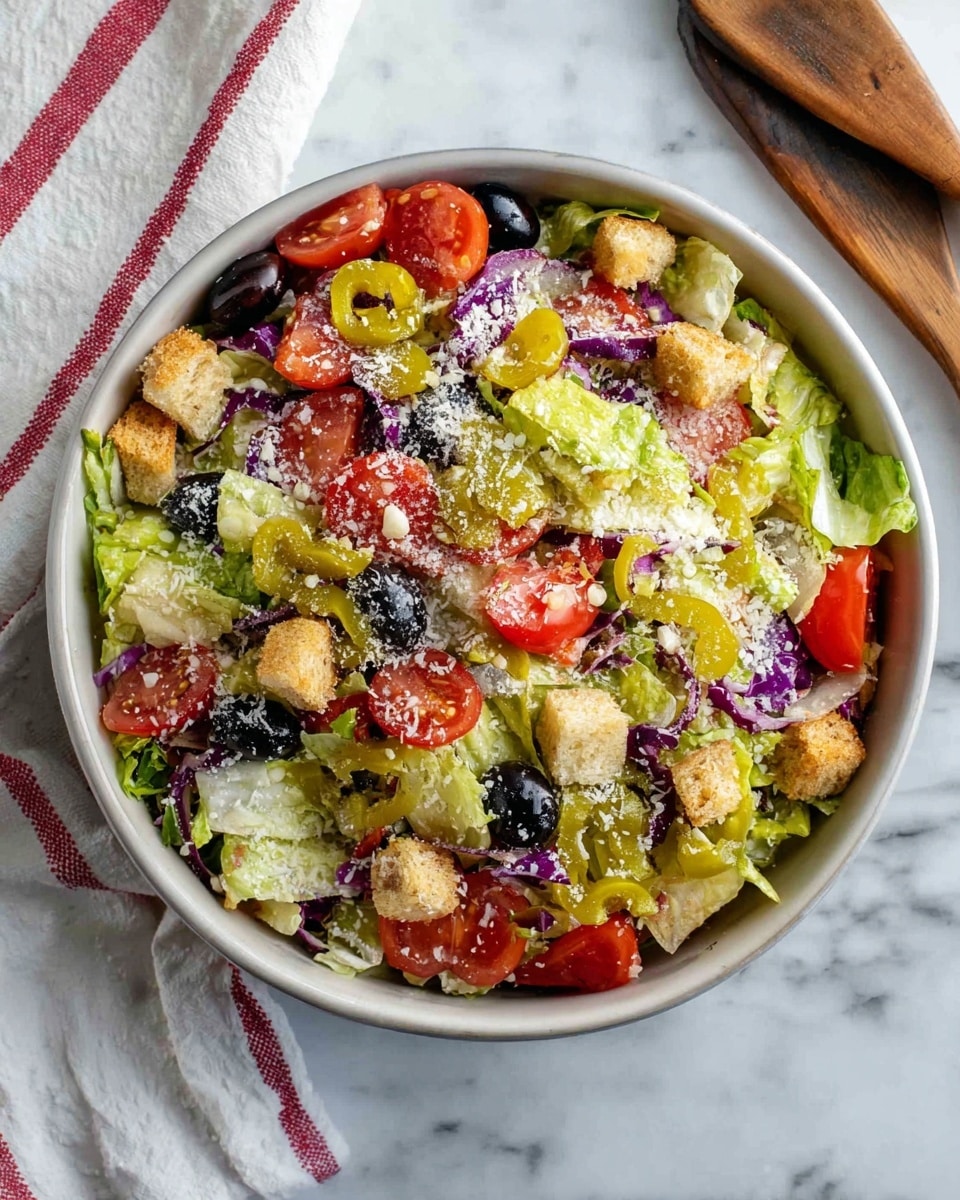 A white bowl filled with a colorful salad sits on a white marbled surface, with wooden salad tongs visible at the top right. The salad has a base layer of green chopped lettuce mixed with small pieces of purple cabbage, topped with slices of red tomatoes and whole black olives. Scattered on top are light brown croutons and several bright yellow pepperoncini peppers. The salad is sprinkled with finely grated white cheese, adding a dusting of texture. Next to the bowl, a white cloth with red stripes is partially visible. Photo taken with an iphone --ar 4:5 --v 7