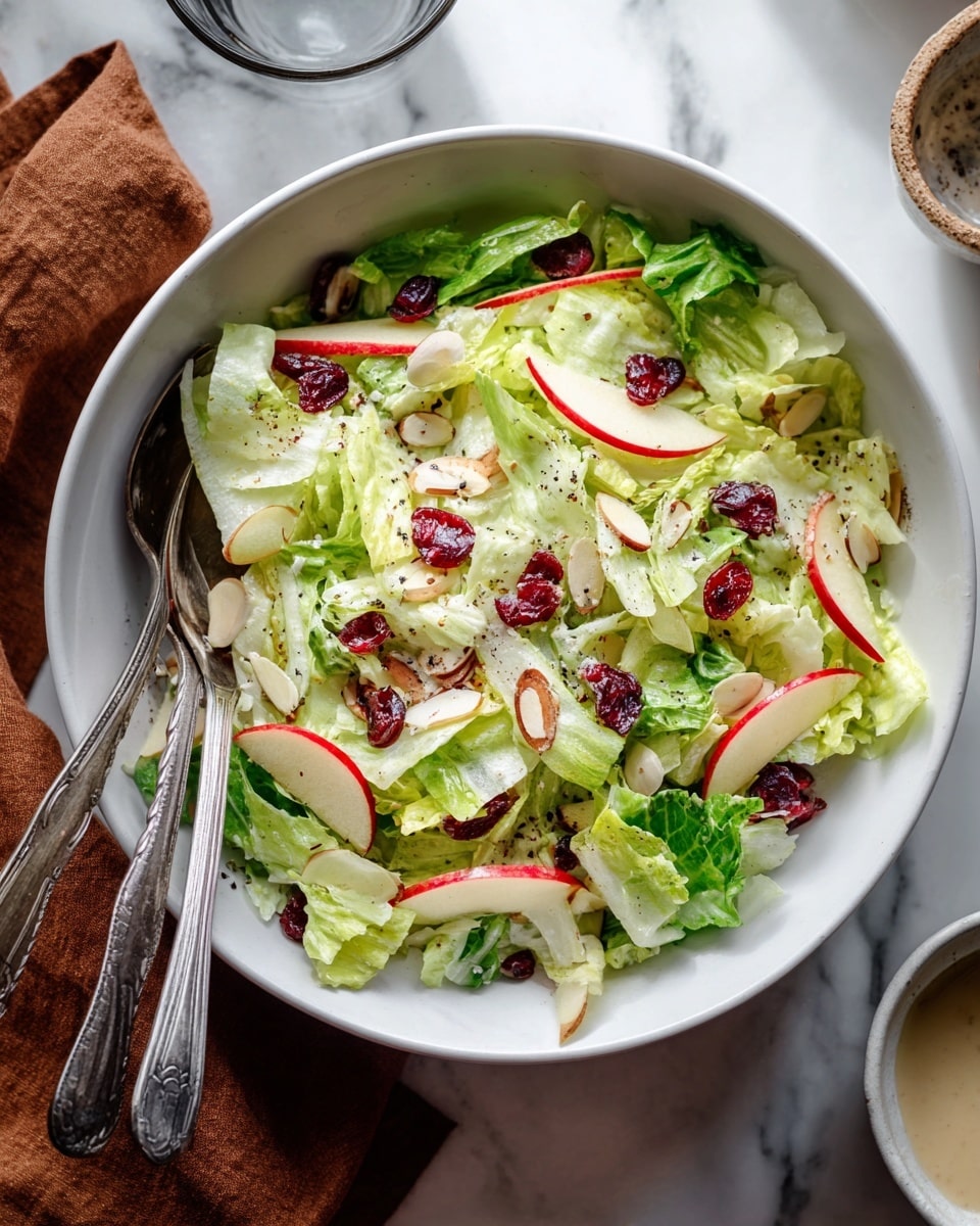 A white bowl filled with a fresh salad made of thinly sliced green lettuce as the base layer, topped with thin red apple slices and scattered red dried cranberries. There are also light green thinly sliced vegetables and a few almond slices mixed throughout, with a sprinkle of black pepper on top. Two metal serving spoons are placed inside the bowl on the left side, ready for serving. The bowl is set on a white marbled surface with a brown cloth napkin on the left and a drinking glass and small bowl of dressing on the right. photo taken with an iphone --ar 4:5 --v 7