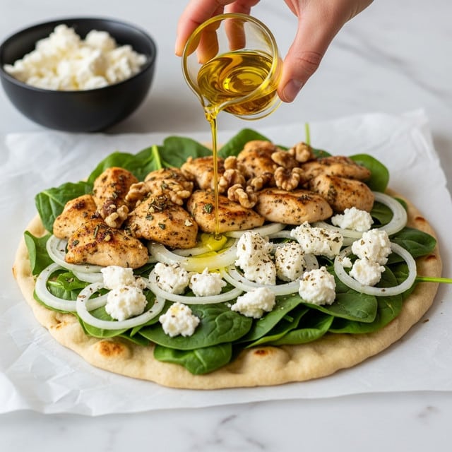 A white flatbread is topped with three main layers: a base layer of green spinach leaves, a middle layer of white onion slices and small chunks of white cheese, and a top layer of golden brown grilled chicken pieces sprinkled with herbs and small bits of walnuts. A woman's hand is pouring a light yellow oil over the flatbread using a small glass container. The flatbread is placed on parchment paper, and there is a black bowl filled with white cheese in the background on a white marbled textured surface. photo taken with an iphone --ar 4:5 --v 7