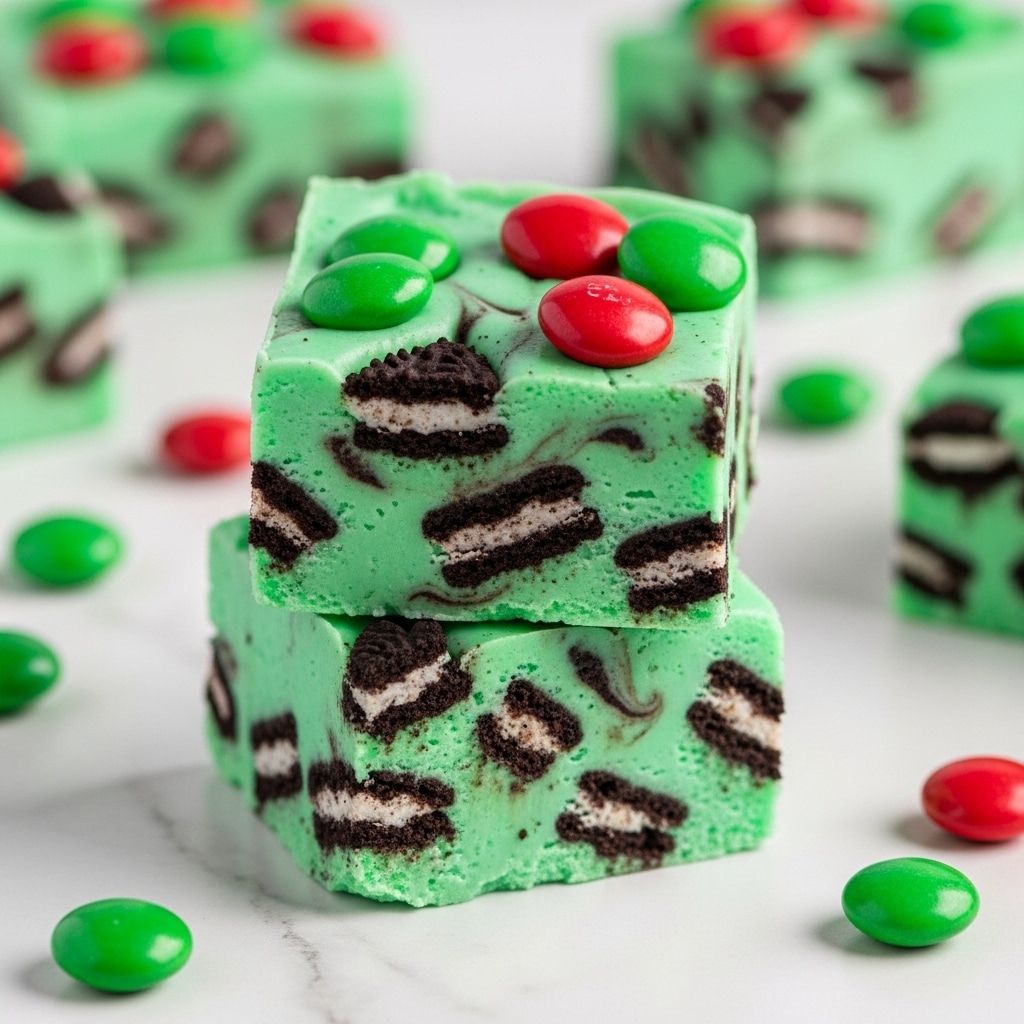 The image shows two square-shaped pieces of green fudge stacked on top of each other on a white marble surface. The fudge has visible dark brown cookie chunks mixed evenly within the bright green creamy base, giving a marbled effect. On the top piece, there are red and green candy-coated chocolates scattered for decoration. A few more pieces of fudge and candy chocolates appear softly blurred in the background. The overall look is colorful and festive. Photo taken with an iphone --ar 4:5 --v 7